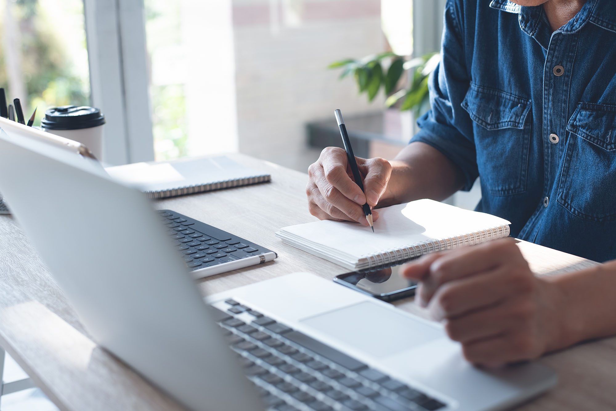 Person in denim shirt taking notes with a pen in a spiral-bound notebook, sitting at a wooden desk with a laptop and smartphone.