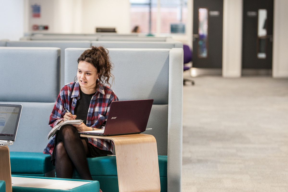 Female student sitting on a sofa in front of a laptop on a small table holding a pen and notebook looking to the side to speak to another person