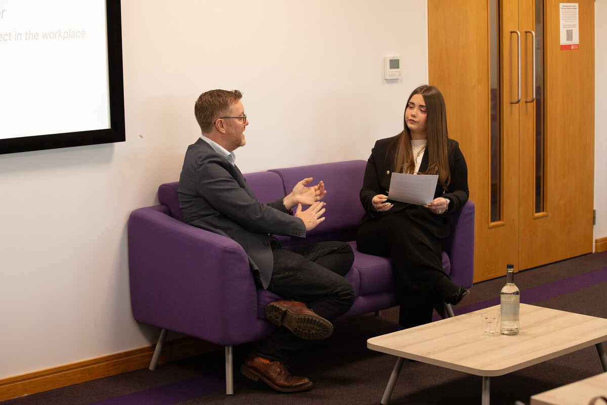Abigail, a student, sat next to guest speaker Tom Sherry in a classroom setting on a purple sofa.