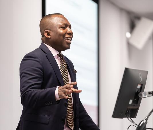 A tutor standing in front of a computer, desk and wall screen, giving a lecture in classroom.