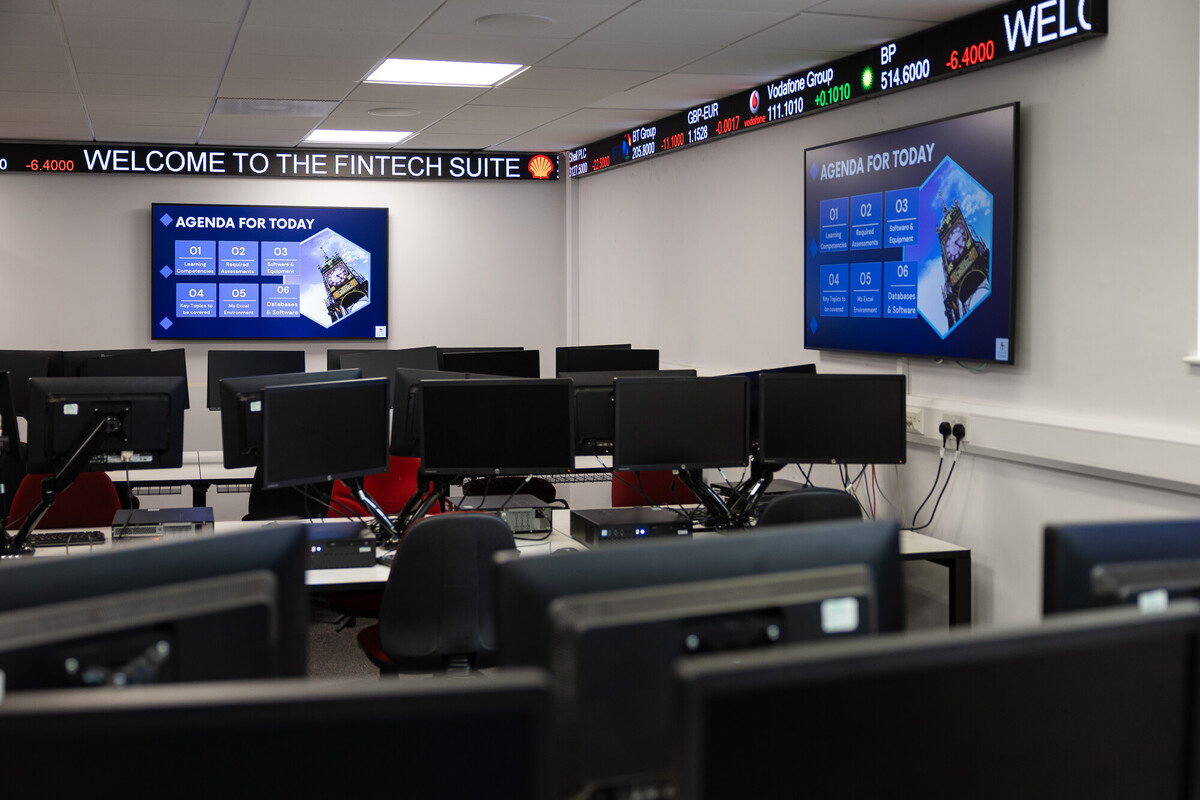 Modern fintech training suite with rows of computer workstations facing large wall displays showing an agenda, beneath a digital stock ticker displaying company prices.