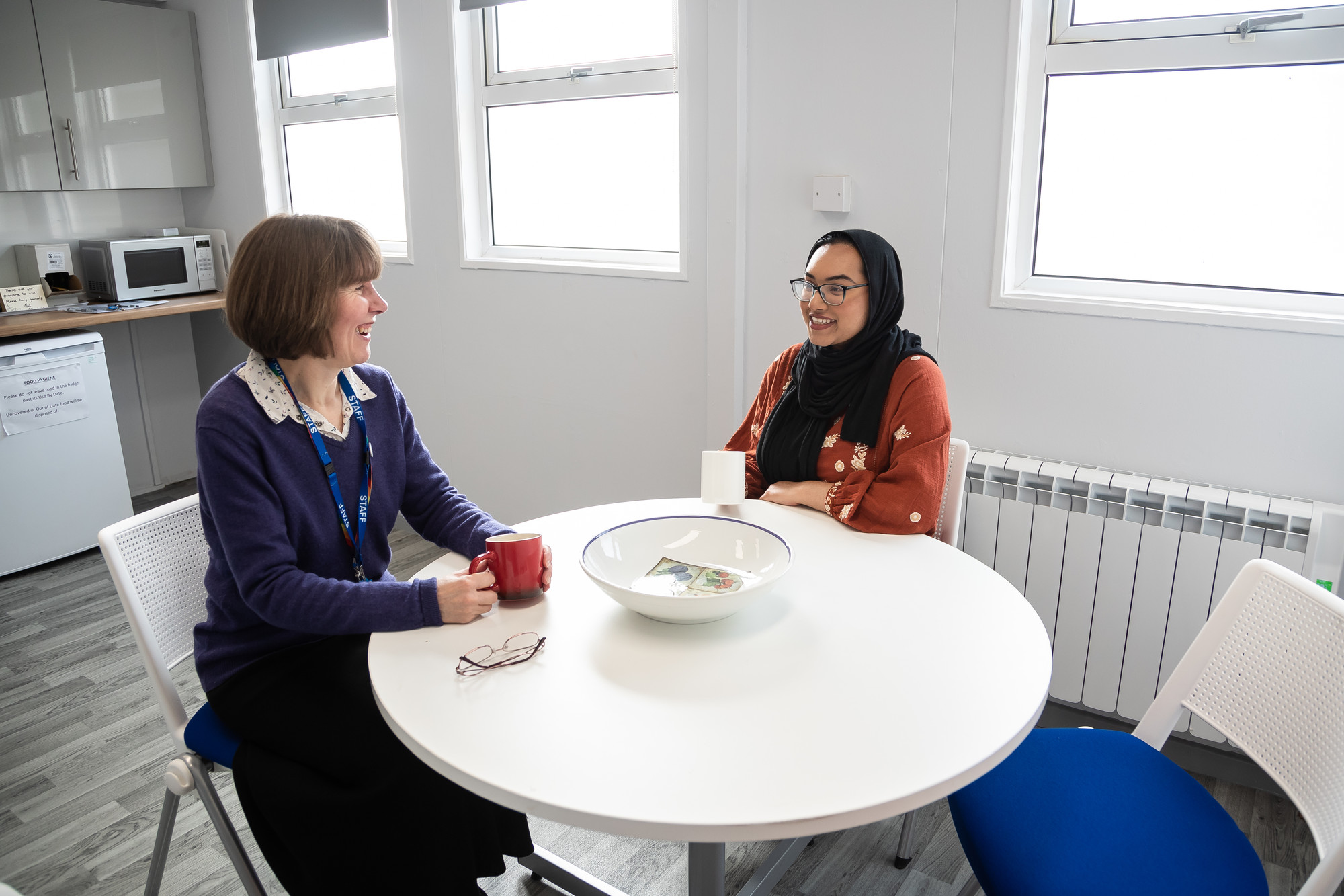 Two people having a casual conversation over coffee at a round white table in a bright modern kitchen with large windows and contemporary appliances.