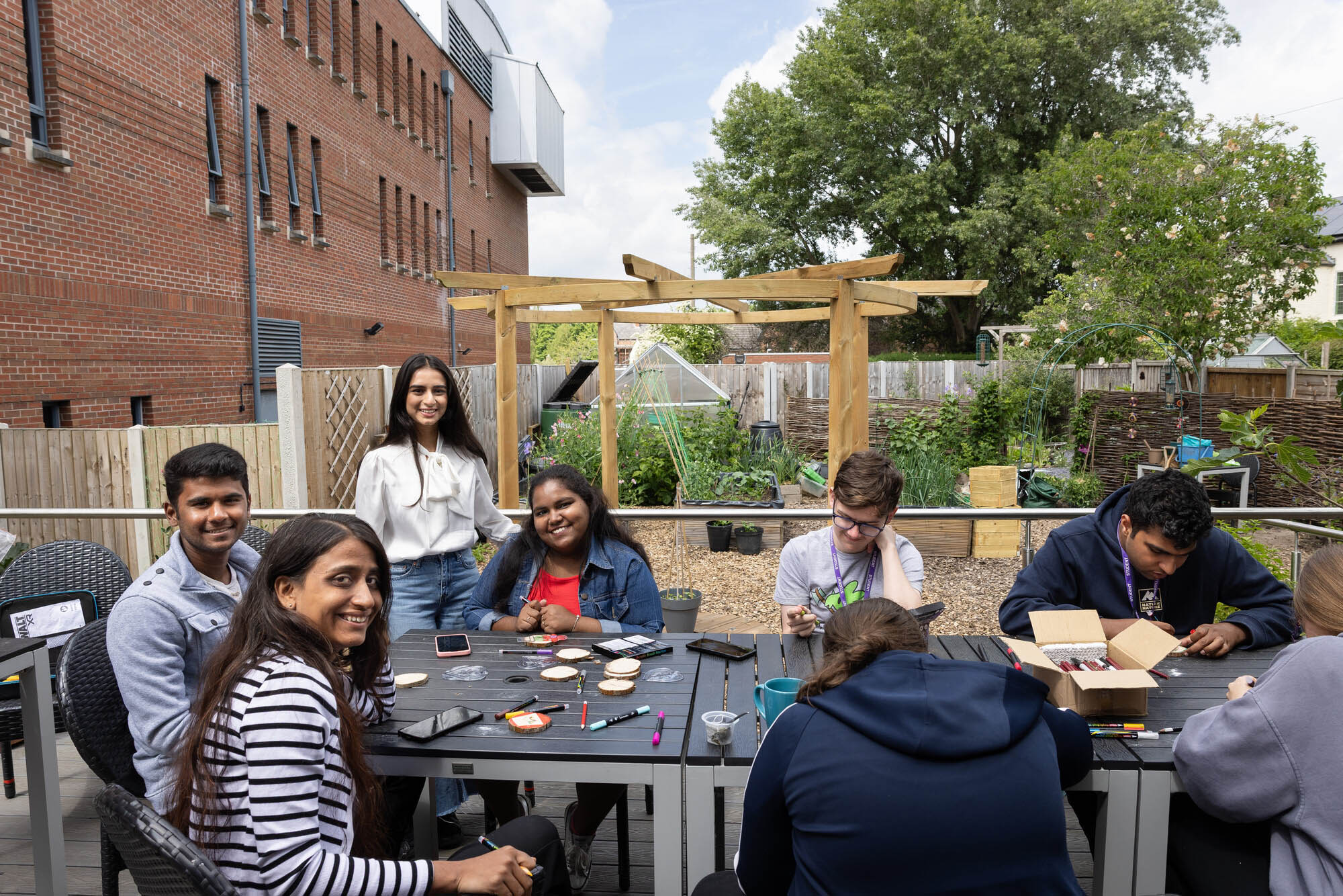 Group of people gathered around an outdoor table under a wooden pergola, engaged in collaborative activities with markers, cups, and devices in a garden setting.