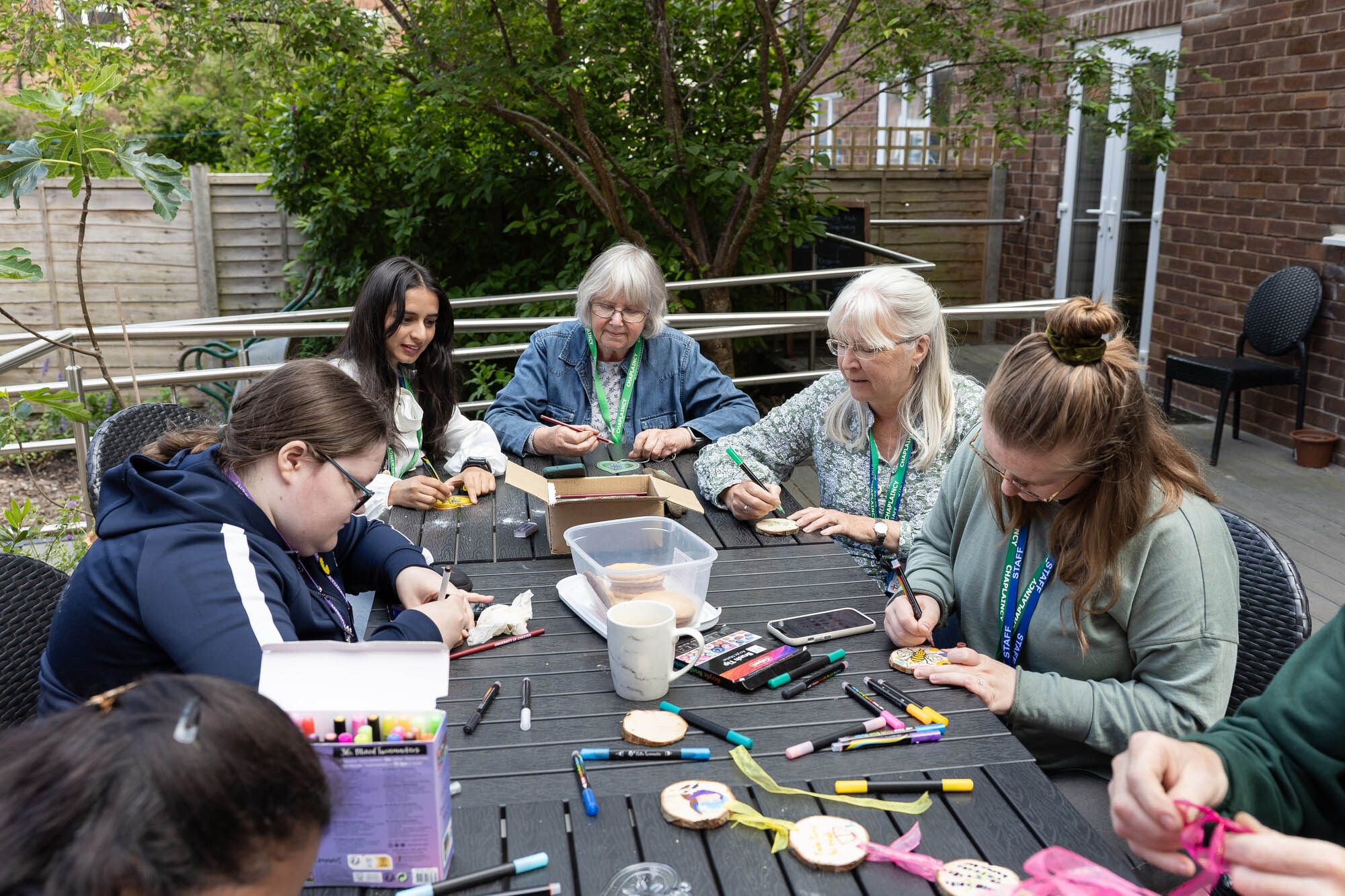 Group of six people engaged in arts and crafts around a black outdoor table covered with markers, wooden discs, and colorful supplies in a garden setting.
