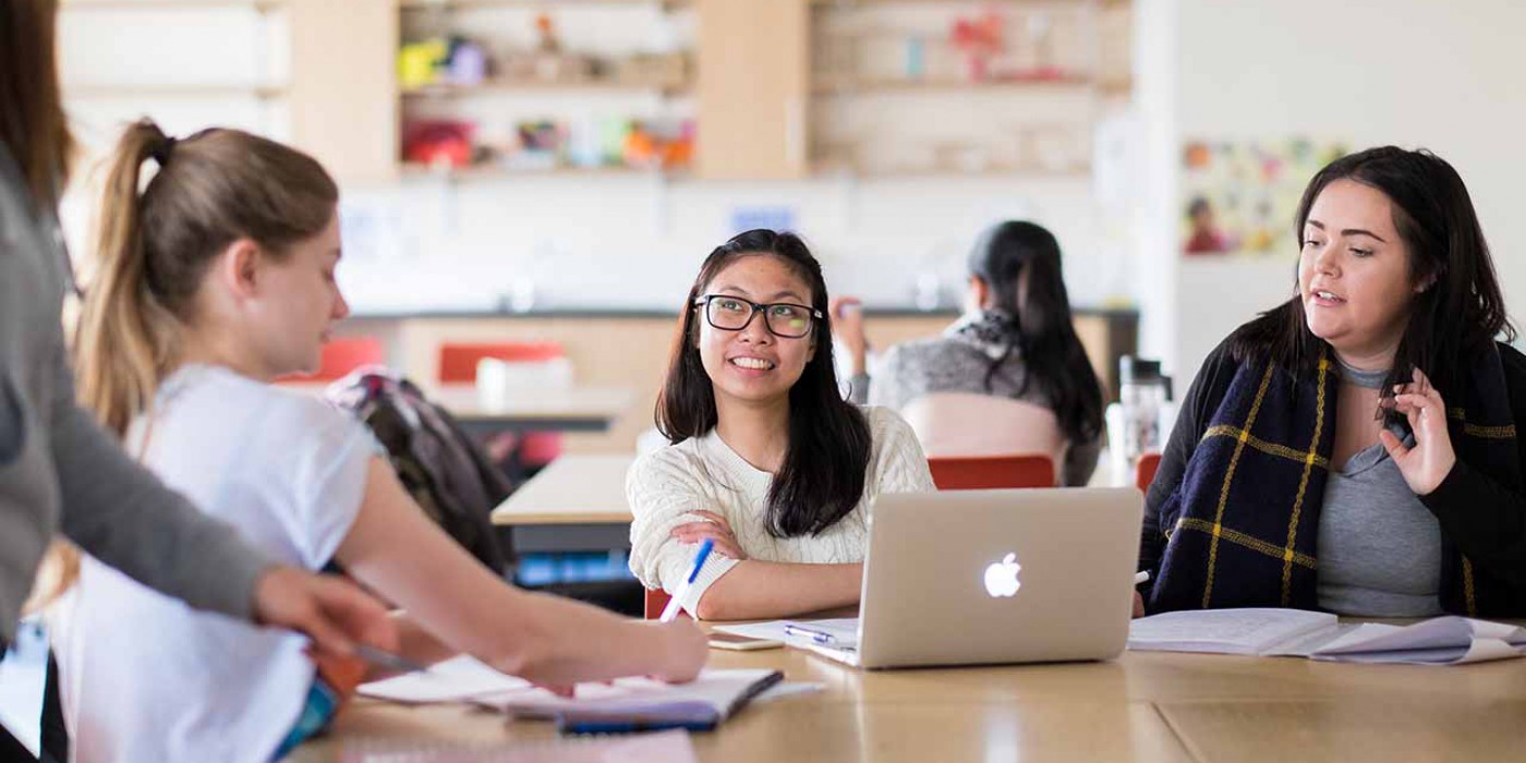 Five students in a classroom, two students facing towards the camera, sitting at a desk together, a third student sits at an angle towards the back of the camera, fourth student stands toward the side and the fifth student is sat at a separate table with her back towards the camera.