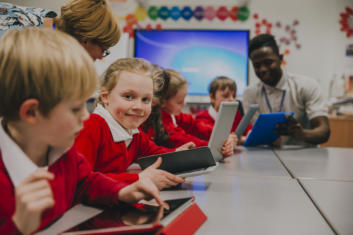 primary school children wearing red school jumpers sitting in a classroom using laptops and tablet devices