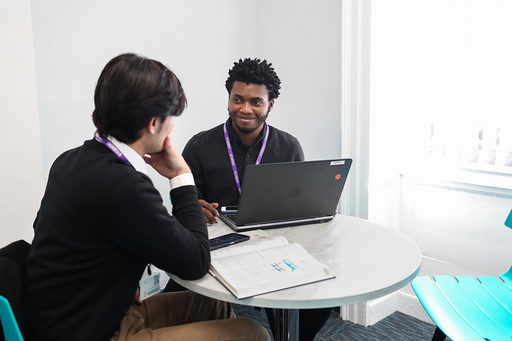 A tutor and student chatting at table with notebook open.