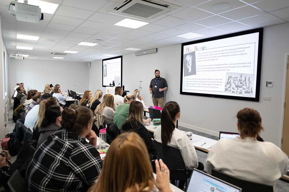 A tutor standing in front of wall screen teaching a class of students.