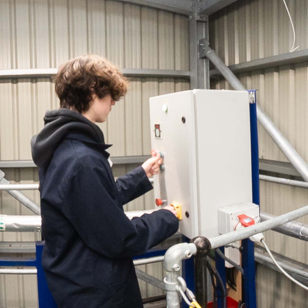 A student in protective outerwear operates a control panel on industrial equipment in a workshop setting.
