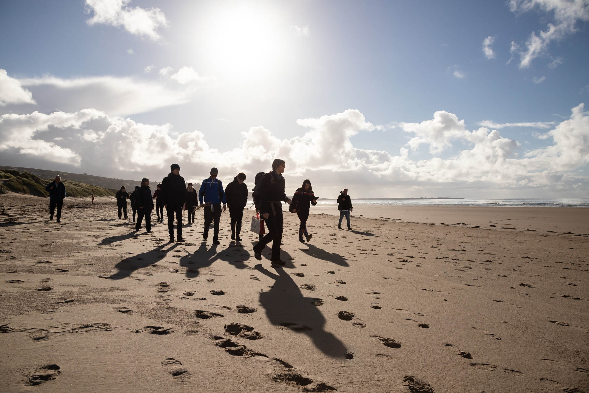 Group of people walking on a sandy beach under a bright sky with long shadows, ocean view, and coastal greenery in the background.