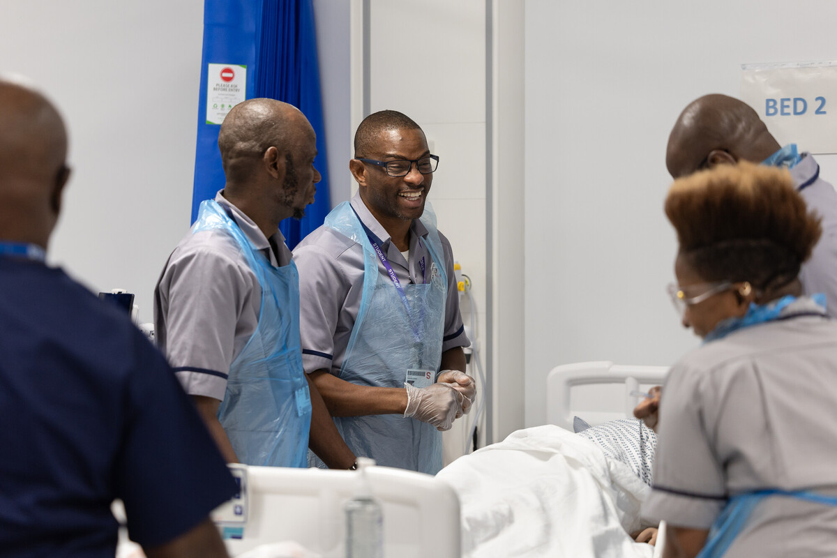 A group of healthcare workers in uniforms and protective aprons gathered around a patient’s hospital bed.