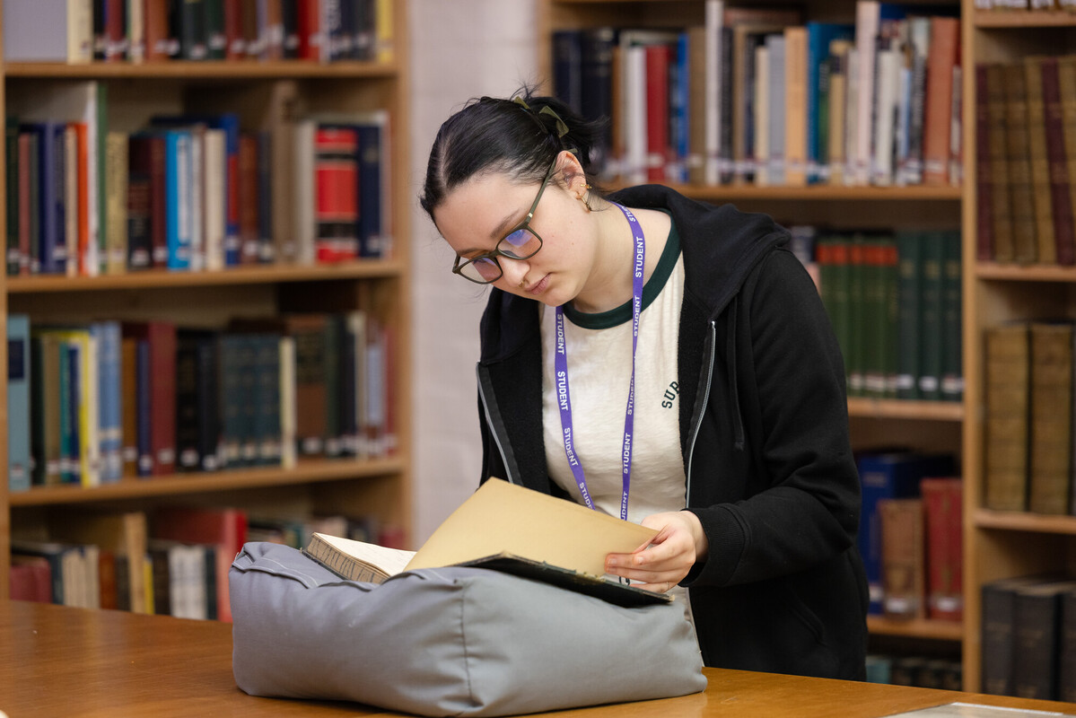 Student examining archival documents in a university library, surrounded by shelves of historical books, highlighting hands-on research and primary source analysis for a History BA degree.
