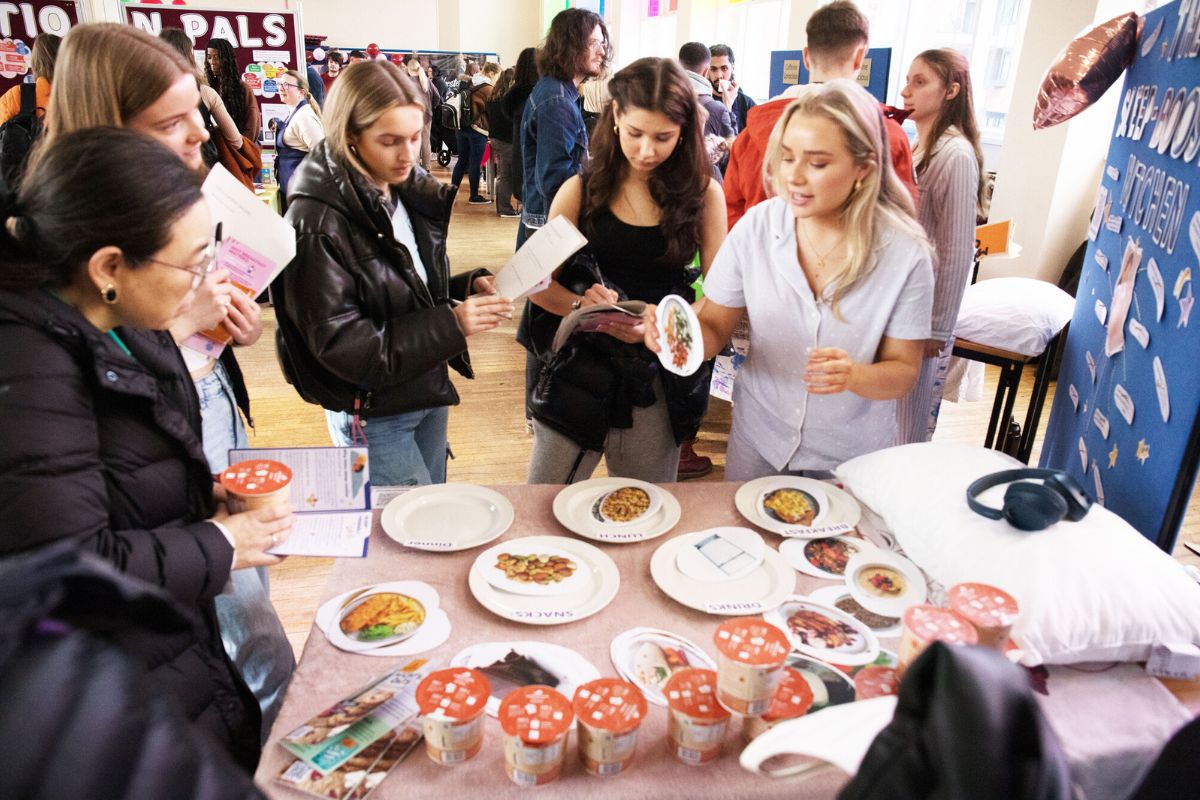A group of students listen to a talk given at The Sleep-Boost Kitchen stall, which includes a table with plates of different labels, leaflets, and snack pots, as well as a pillow and headphones.