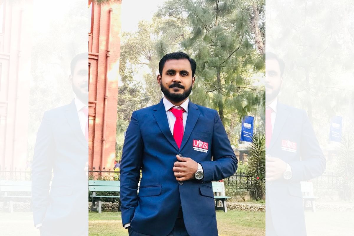 A man in a blue suit and a red tie facing the camera. The suit as a 'UVAS' logo on it. Trees, benches and a building are in the background.