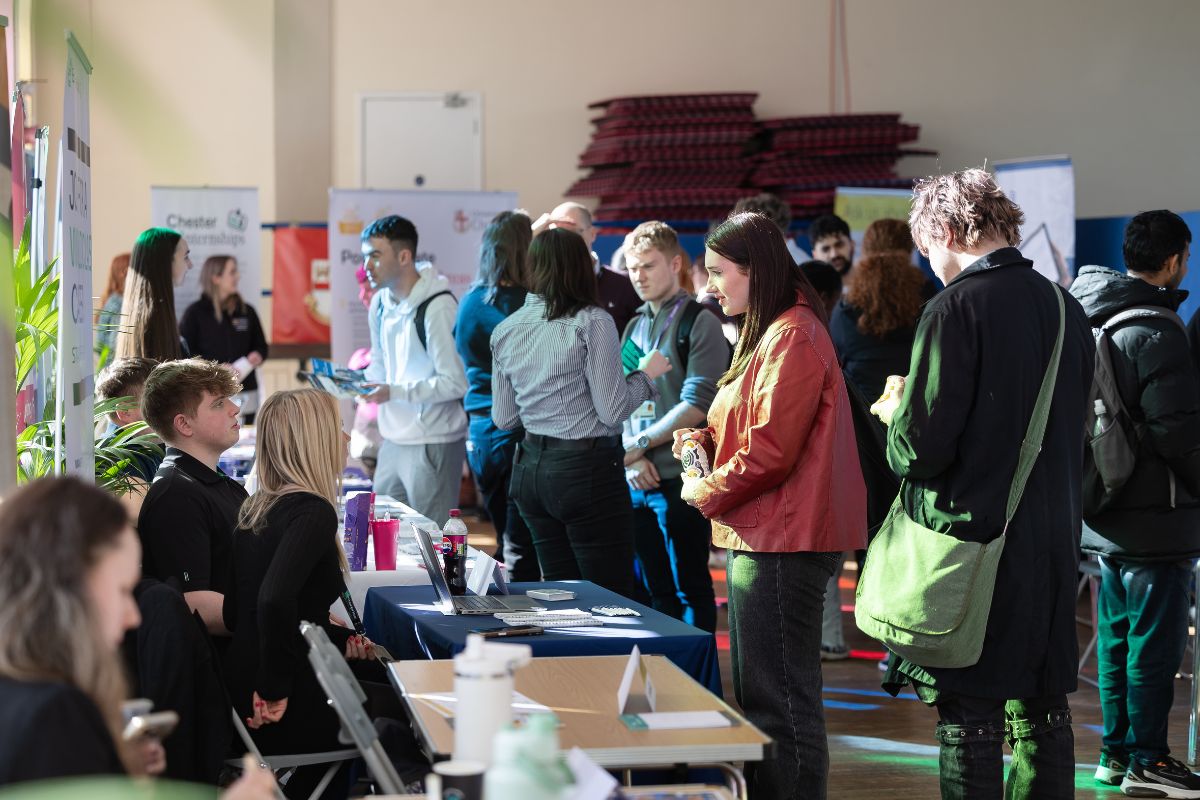 A group of people in a busy room looking at stalls and in discussions with people sat at tables.