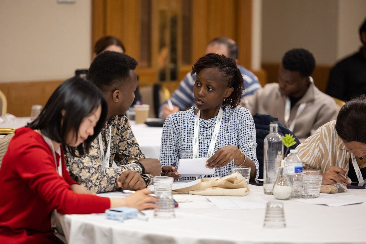 A group of people sat at a table. Two of the people are in a discussion, the other two are looking down at the table, possibly some writing. In the background is another table with people sat around it.