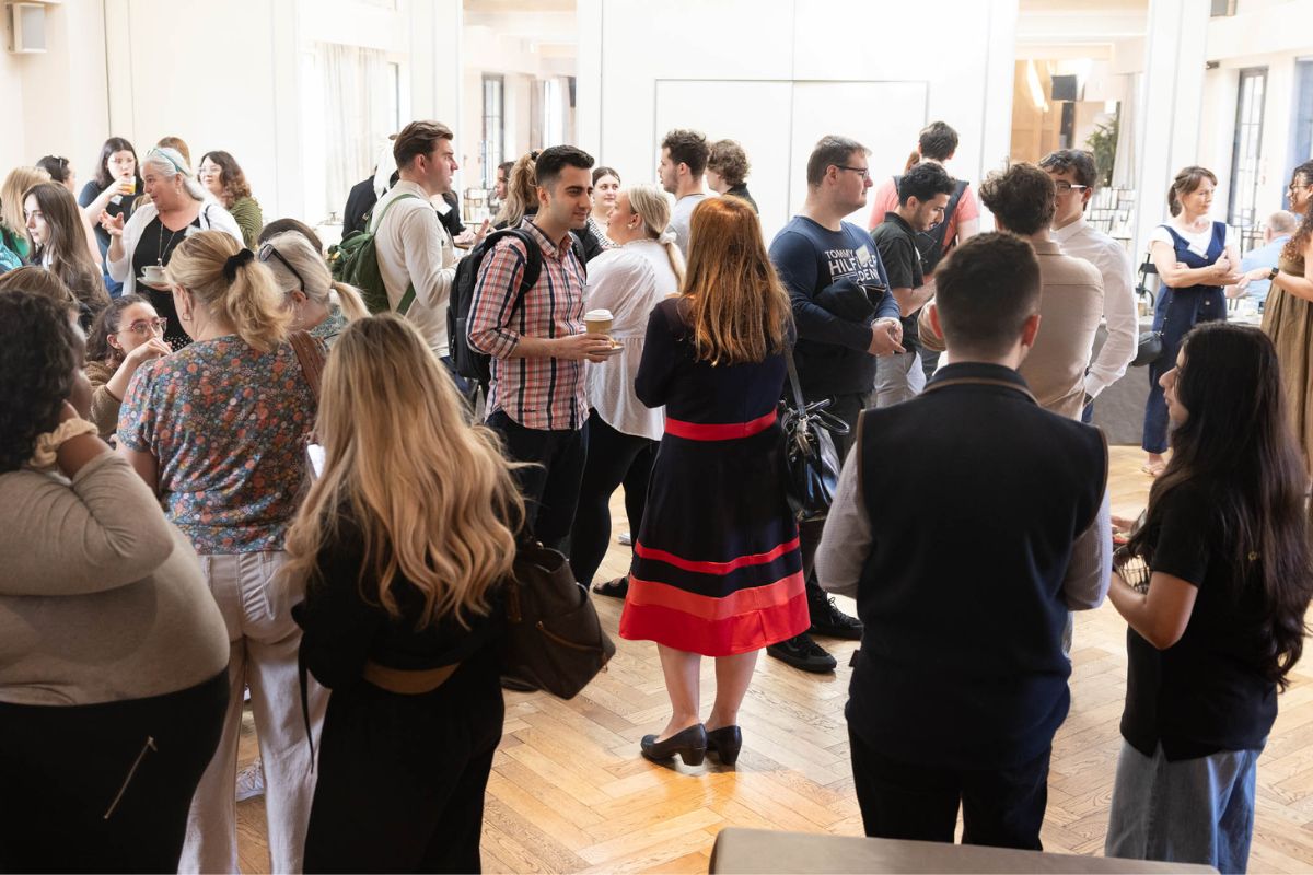 A picture taken from the back of the room showing a group of people in a busy environment talking to each other.