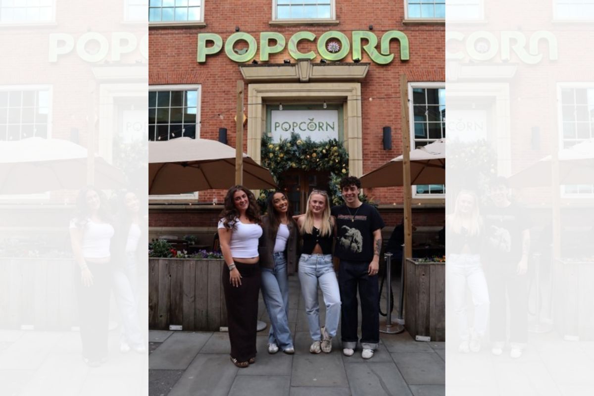 Four students standing, smiling, facing the camera, on the outside of the Popcorn bar. Two big 'Popcorn' signs are visible in the background.