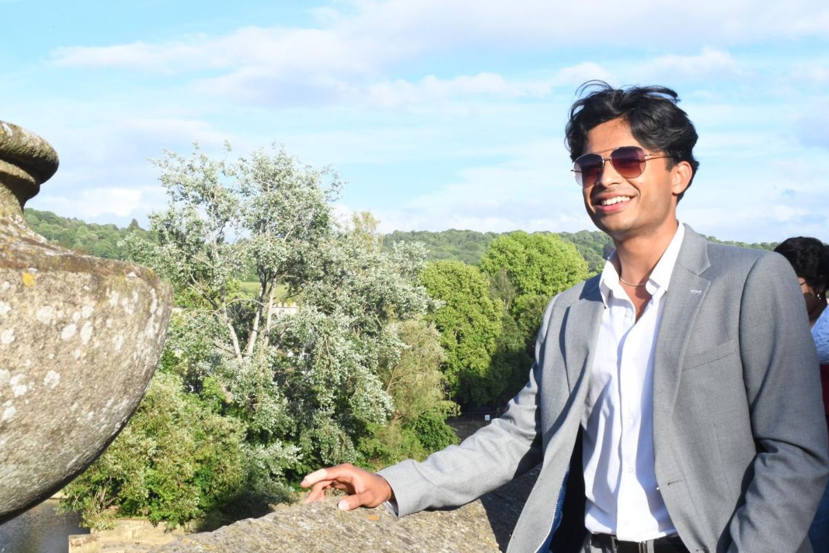 A picture of Aadhi wearing sunglasses, smiling, resting his right hand on a stone wall.