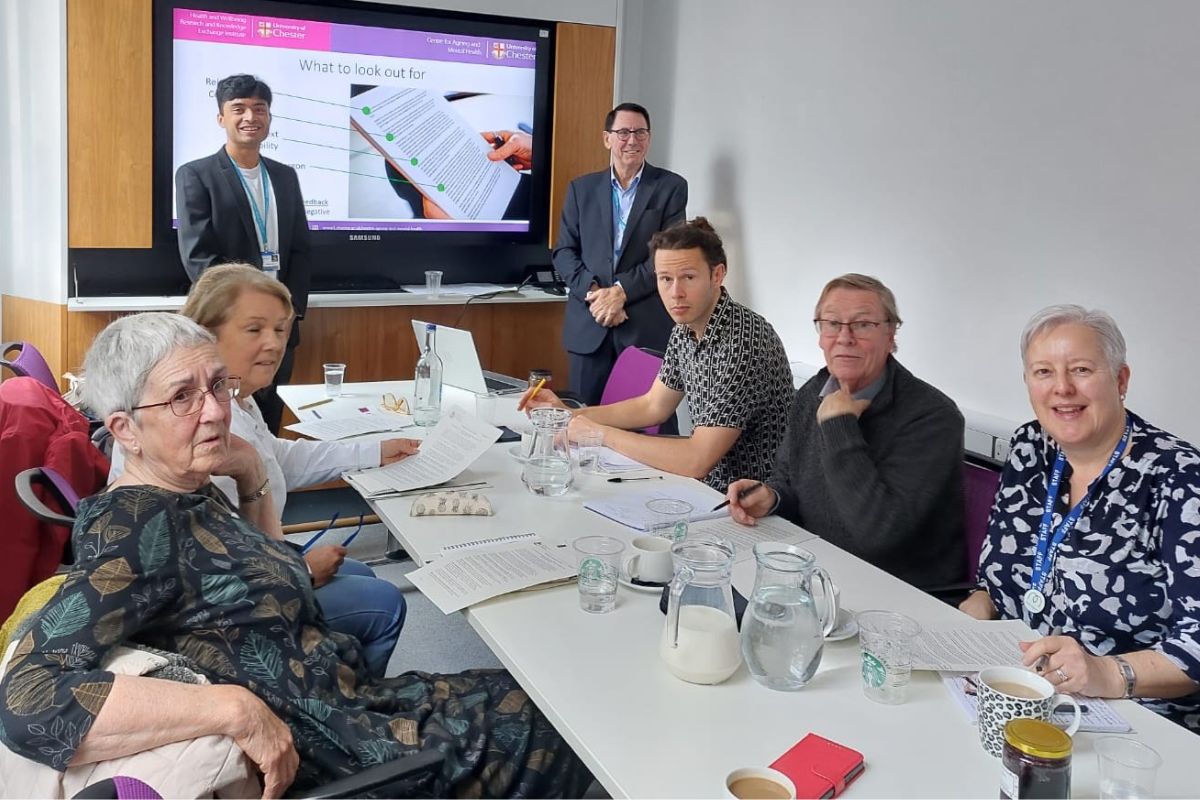 A group of people sat around a table with documents on it. Two people, including Aadhi, are stood at the back of the picture, in front of a screen displaying a lecture slide. Aadhi is looking at the camera, smiling.