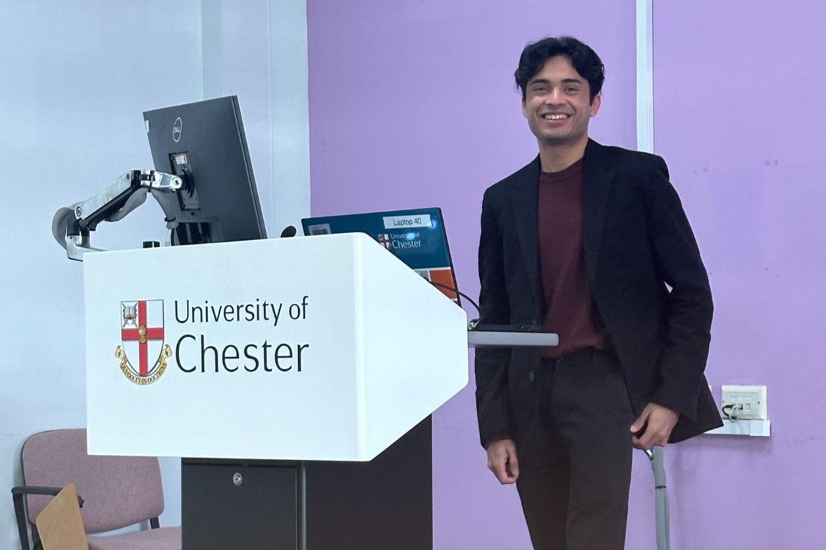 Aadhi Agilan at a lectern which is displaying the University of Chester logo. Aadhi is looking at the camera and smiling.