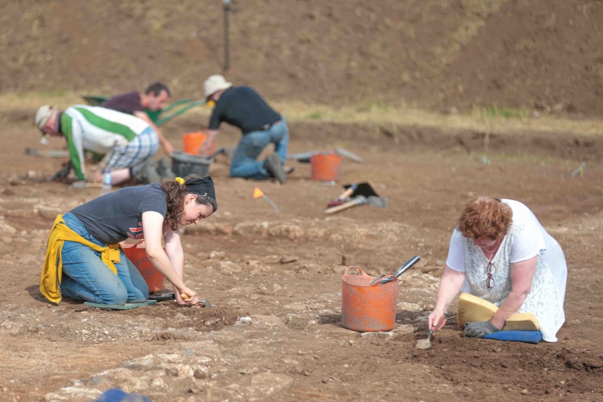 A group of people crouched down at an archaeological dig with trowels and buckets.