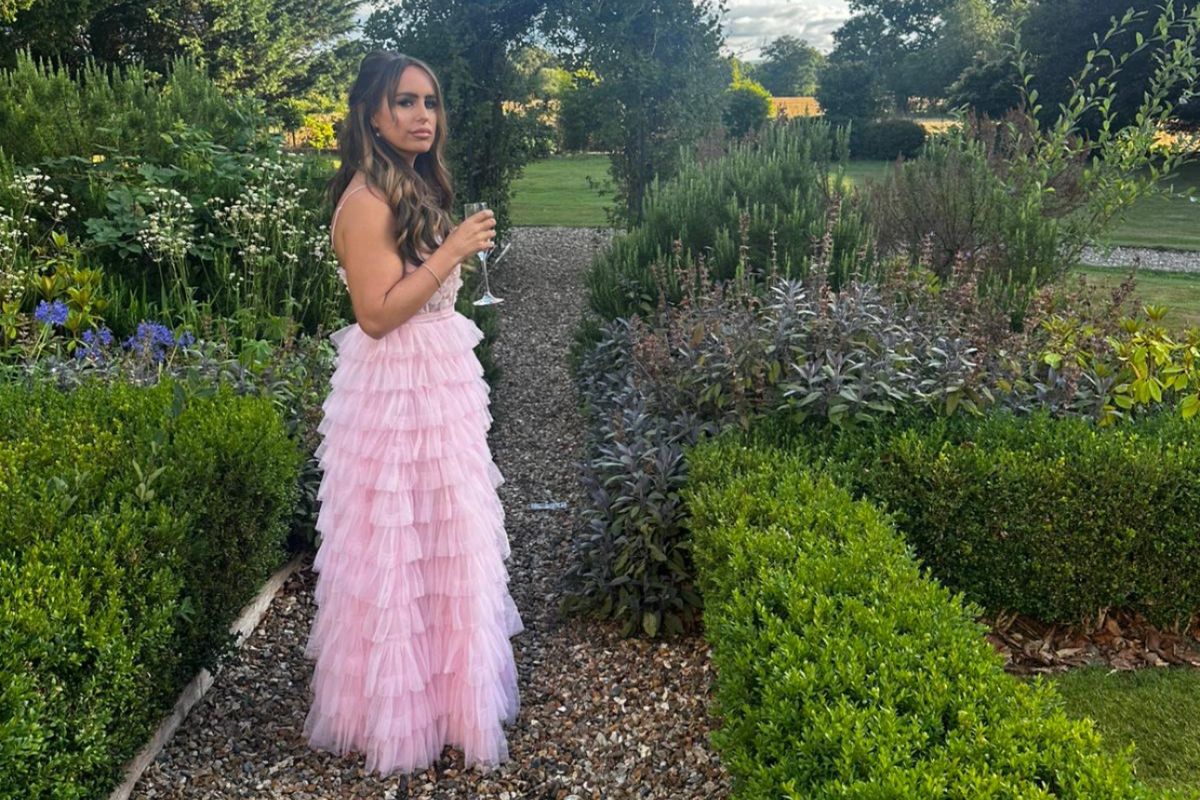 A woman in a pink dress, looking at the camera, holding a glass of drink, while standing in a garden
