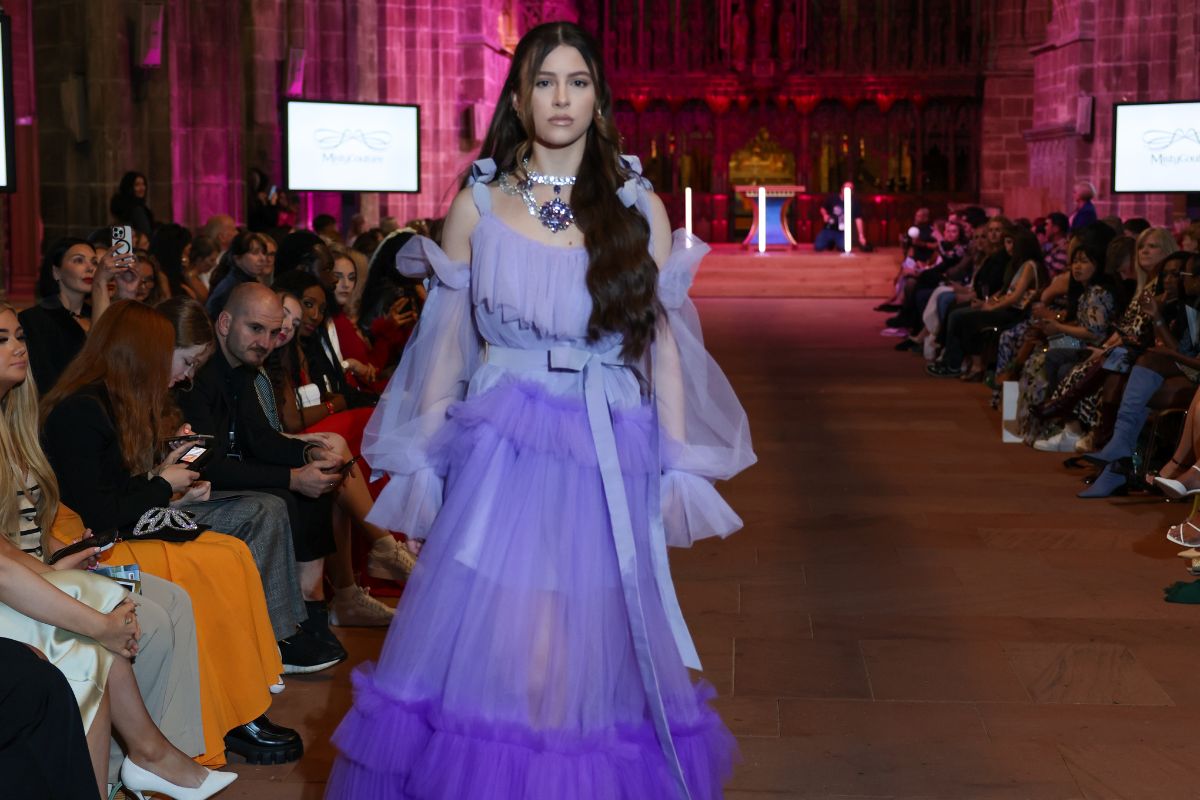A fashion model in a lilac and violet designer outfit walking on a stone catwalk in front of a sitting crowd at Chester Cathedral.