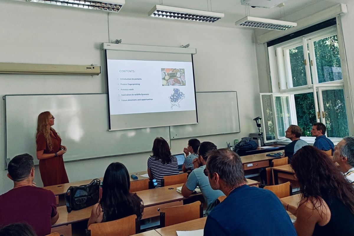 A woman giving a class in a room with people sat down. The screen on display at the front of the classroom includes the words 'introduction to proteins' and 'proteins fingerprinting'.