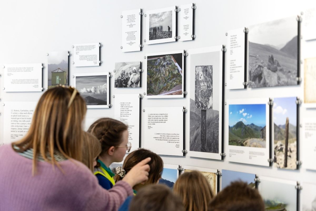 A rear view of people looking at a collection of pictures and words on a wall as part of an exhibition. The group includes schoolboys and schoolgirls, as well as a woman pointing at one of the displays.