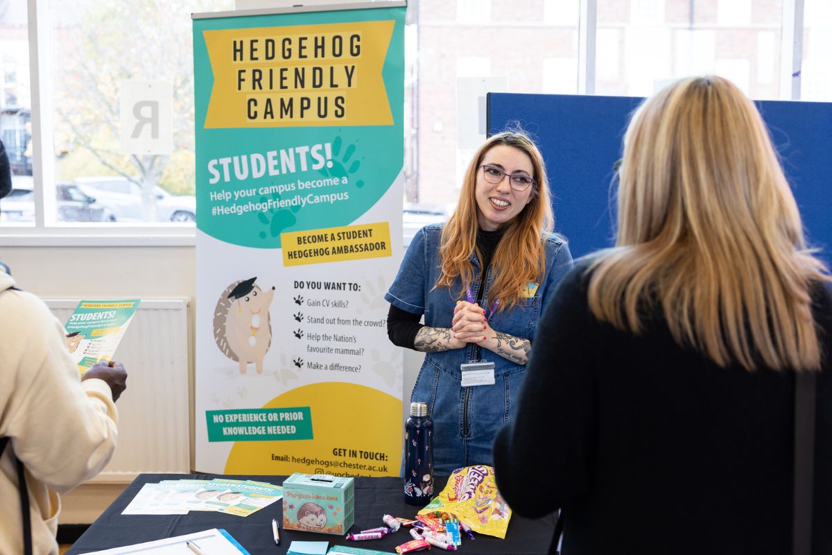 A woman at a stall smiling and looking at an attendee at the fair. The stall has a banner in the background titled 'Hedgehog Friendly Campus' and the message 'Students! Help your campus become a Hedgehog Friendly Campus', with further details.