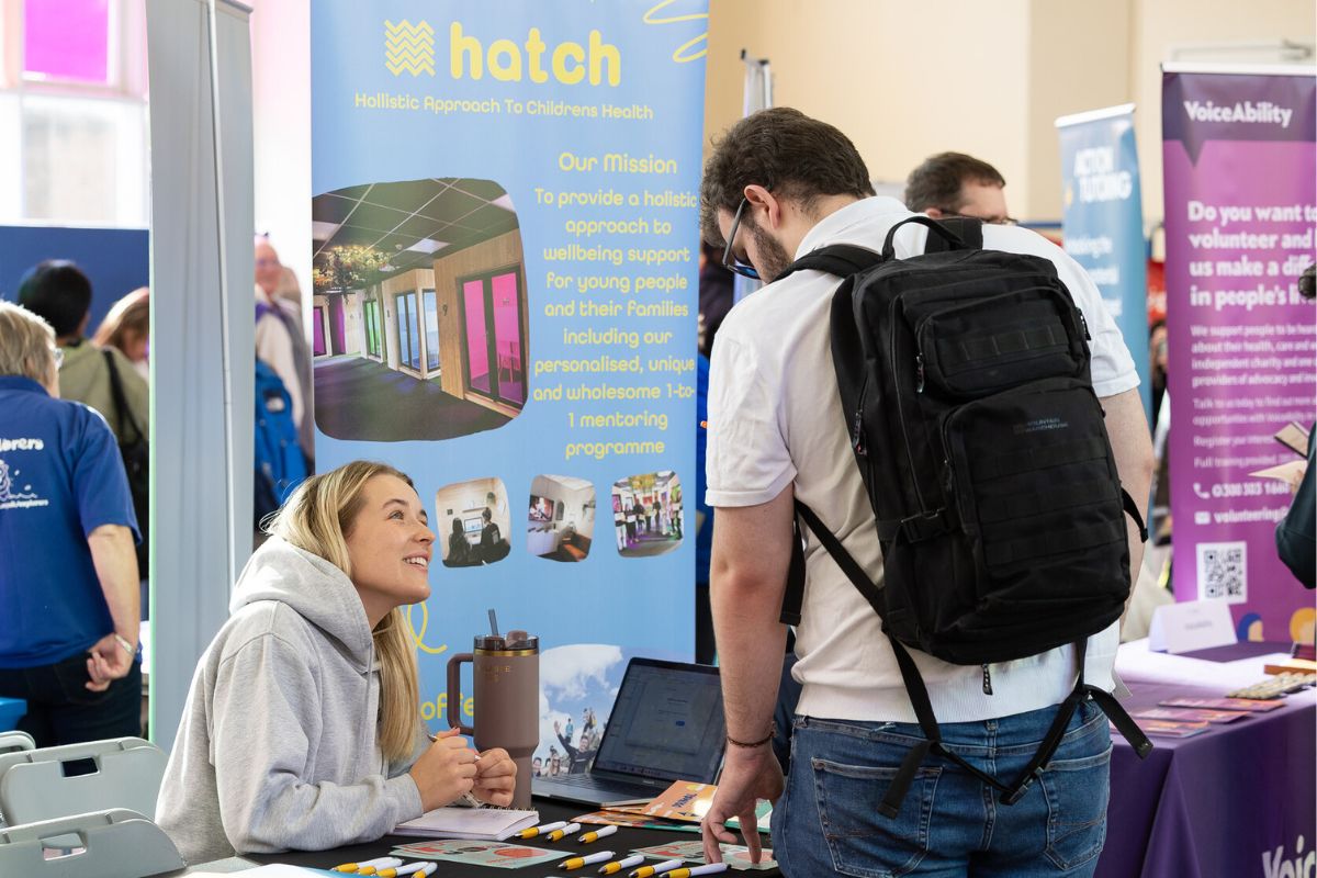 A student with a rucksack on the right leaning over as a stallholder woman, left, looks up at him, smiling. The background information stand is titled 'Hatch: Holistic Approach to Children's Health', with information about the organisation.