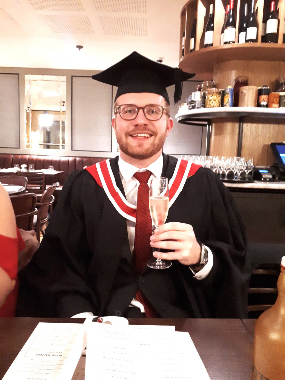 A male student, wearing graduation gown, is holding a glass of champagne