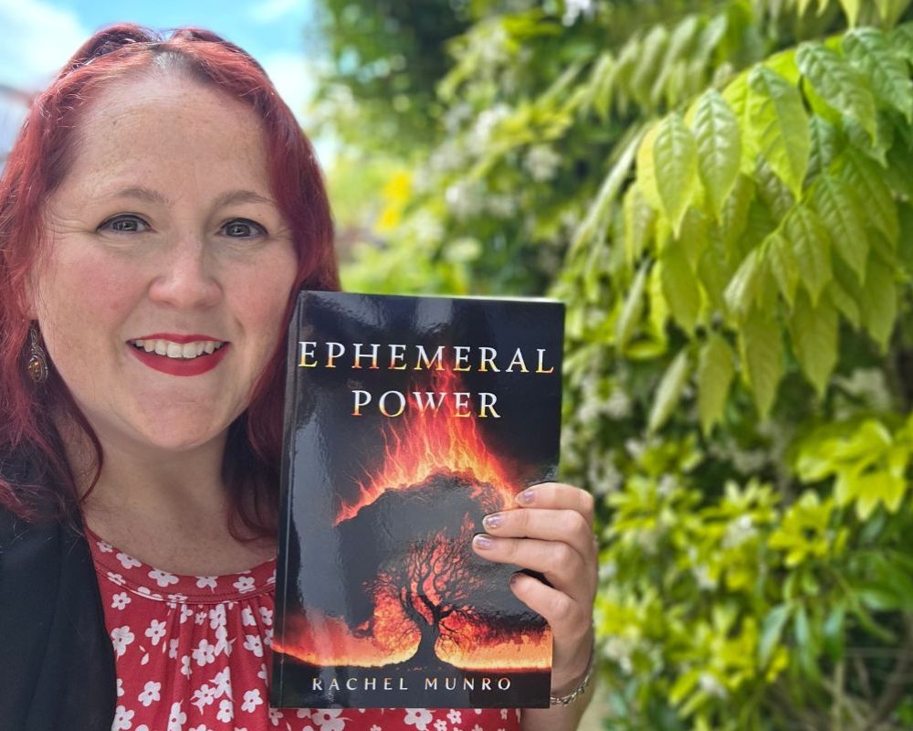 Rachel Munro smiling at the camera while holding a copy of her book, titled Ephemeral Power. The book cover is mostly in silhouette, with flames rising from a tree.
