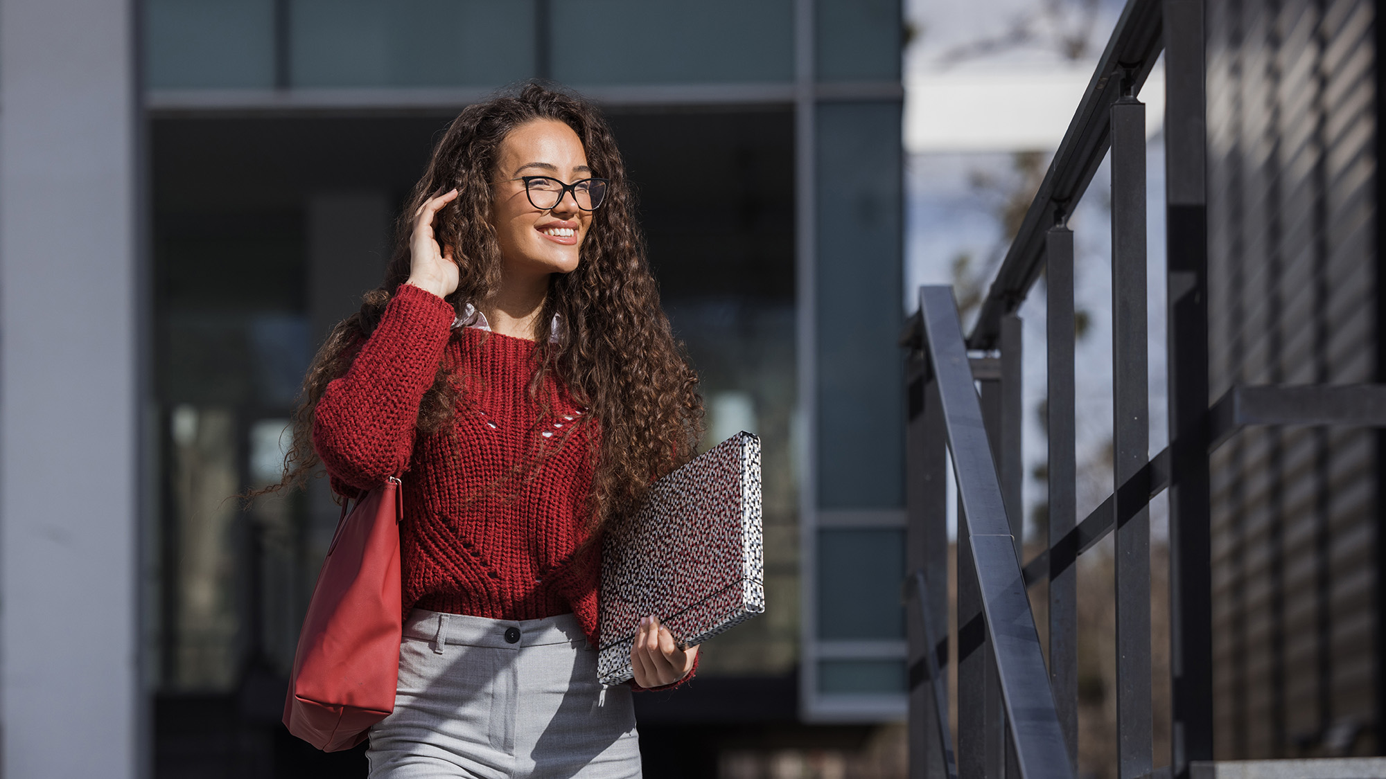 Female student in red top with big smile and one hand in hair while holding a folder with the other.