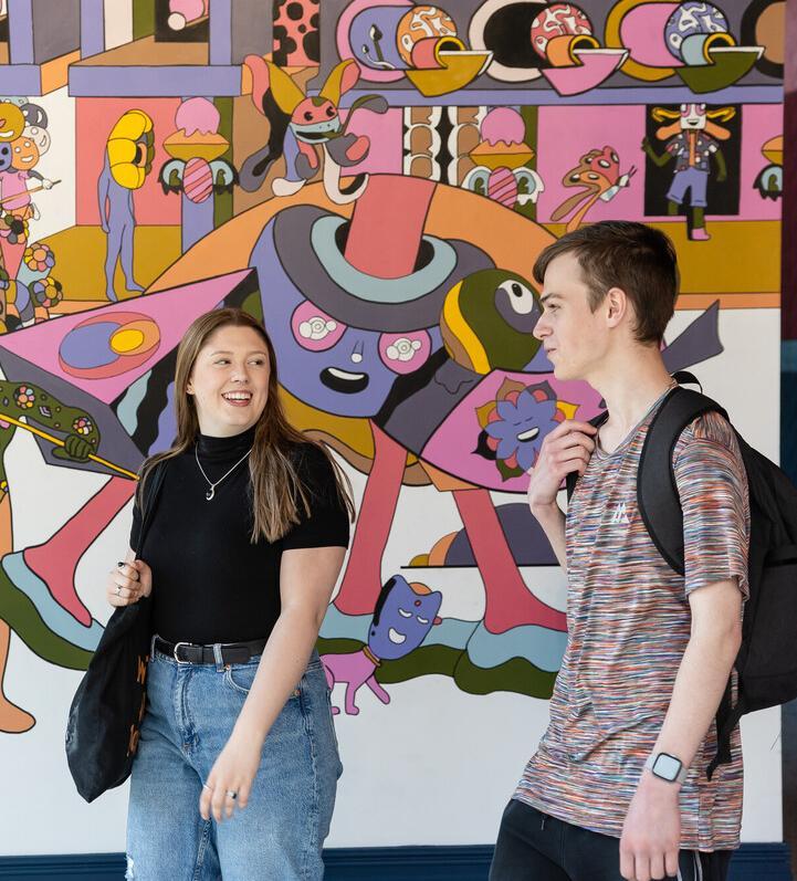 A male and female student, walking and talking in front of a colourful murial wall in Chester.