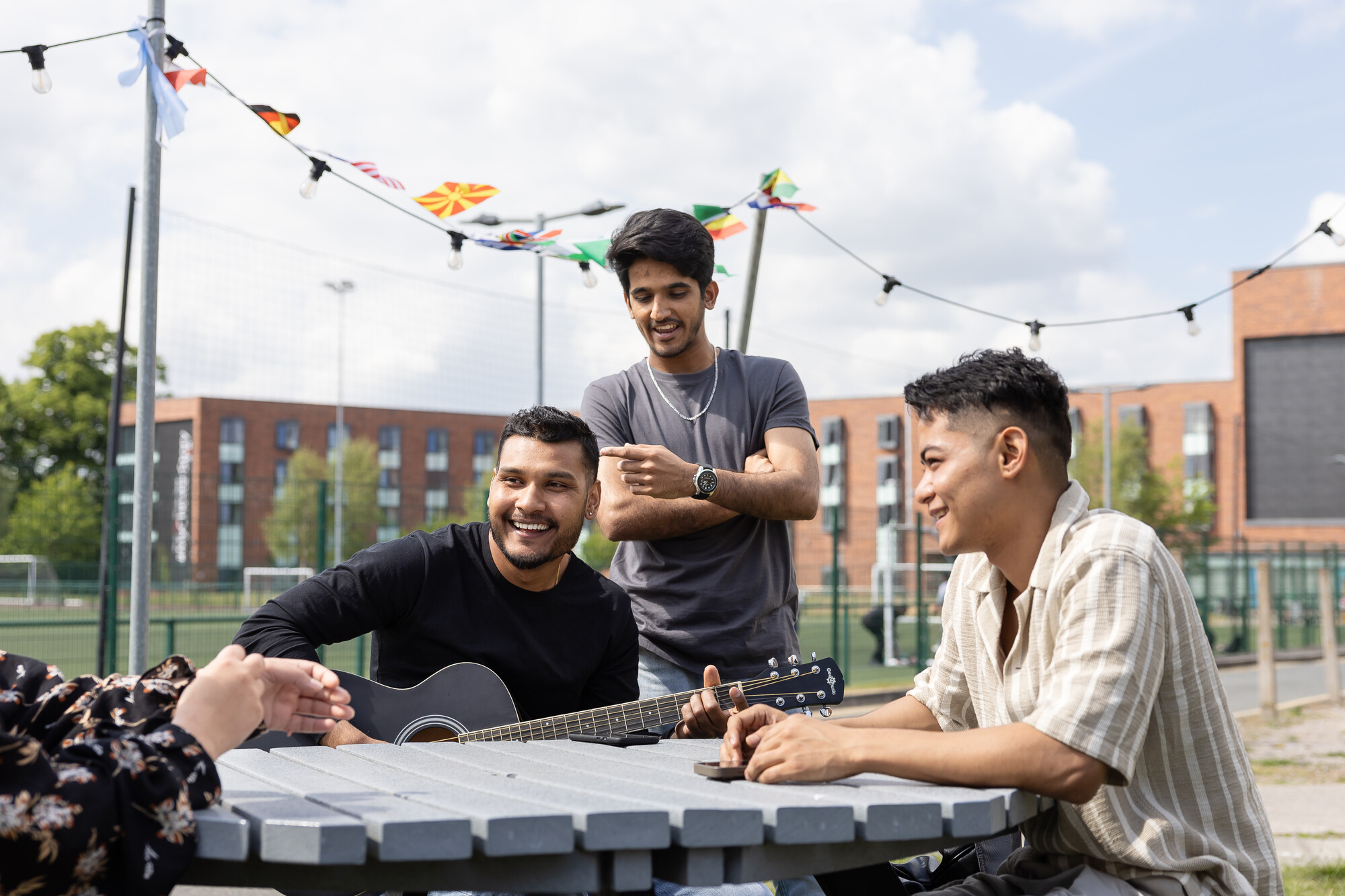 Students enjoying outdoor social activities at the University of Chester campus, sitting around a table with a guitar, festive string lights, and modern accommodation buildings in the background.