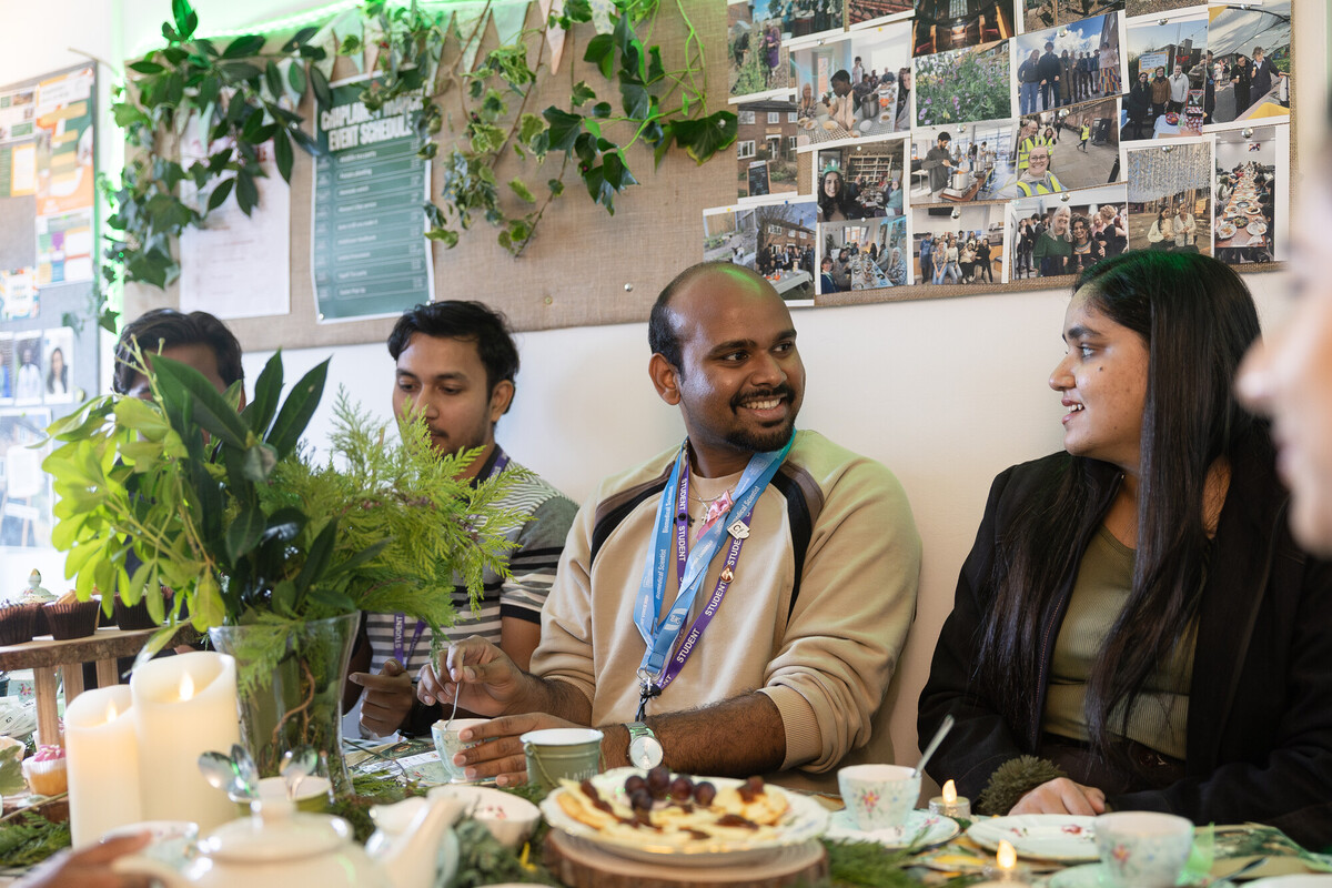 People sitting at a decorated table with plants and candles, sharing tea and desserts in a cosy indoor setting with photos displayed on the wall behind them.